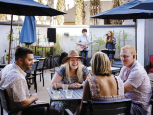 four adults sitting at an outdoor table , smiling with cups on the table with 2 musicians performing in the background