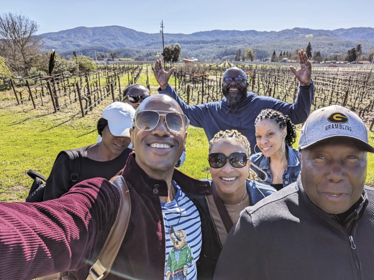group of adults in front of vineyard with mountain and blue sky in the background, smiling taking a selfie