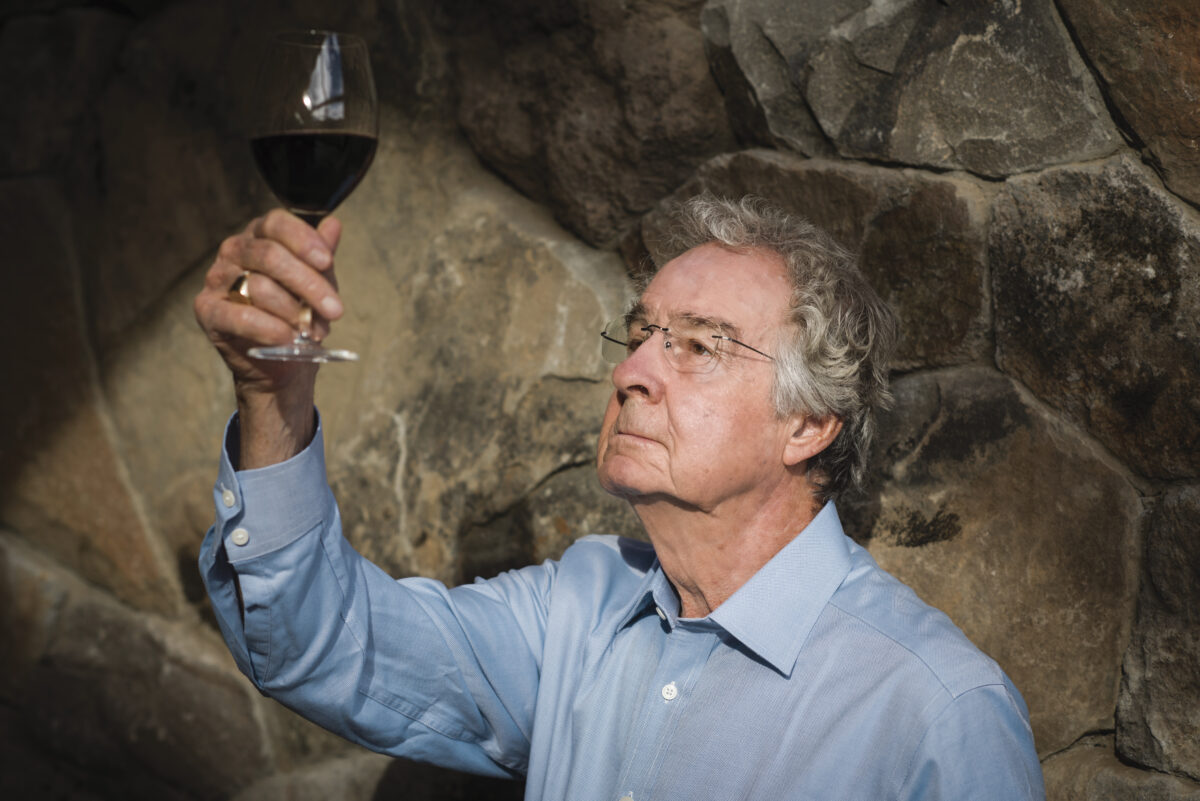 Warren Winiarski wearing a light blue button down shirt and glasses, looking and holding up a glass of red wine