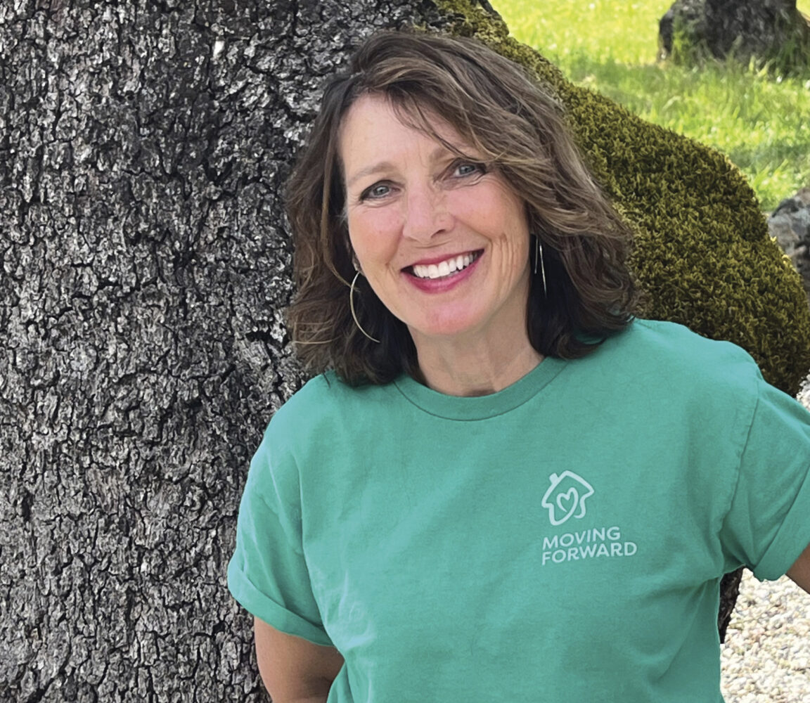 Marita Musante in green "Moving Forward" tshirt standing in front of tree, smiling