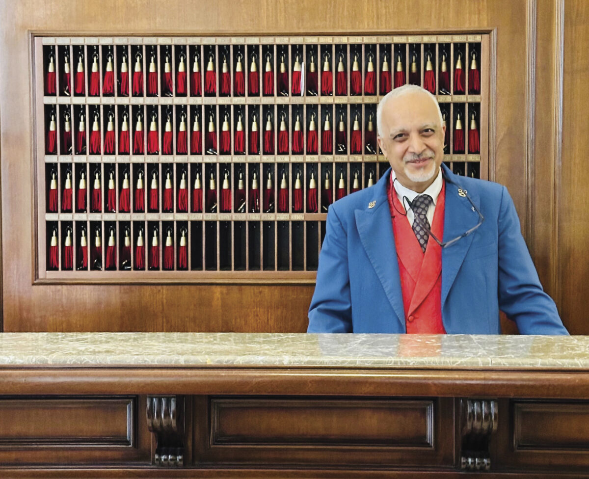 Man in blue blazer and red vest with tie, standing behind hotel desk with keys behind him, smiling