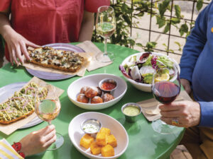 people at table with green tablecloth with a spread of brightly colored food in white bowls including salad and boards with flatbread with glasses of wine in hand