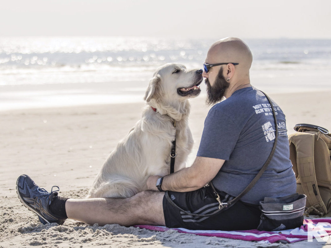 white lab retriever sitting face to face with man with beard wearing sunglasses on beach