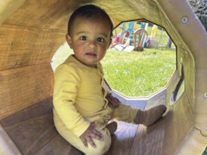 young child in yellow romper in wooden play tunnel with green grass and colorful play area in background