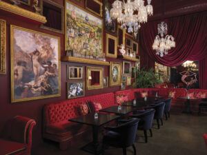 lavish interior with gallery wall of ornate paintings and art with gold frames, red backed boothe with black tables and chairs in red room with chandeliers