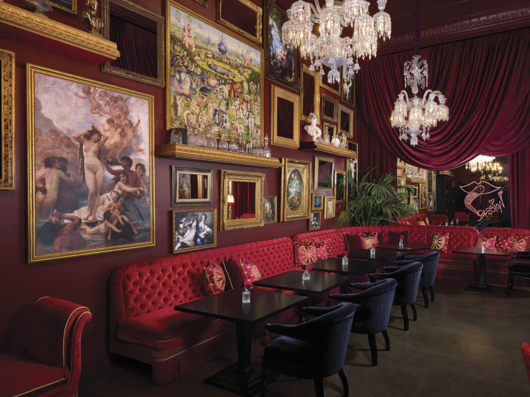 lavish interior with gallery wall of ornate paintings and art with gold frames, red backed boothe with black tables and chairs in red room with chandeliers