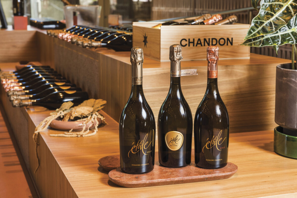 display of 3 bottles of Chandon wine on wood tray on counter with wine and boxes in background