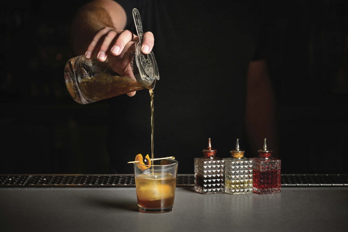 person pouring a cocktail from a shaker into a glass with 3 other cocktails to the side with dark background