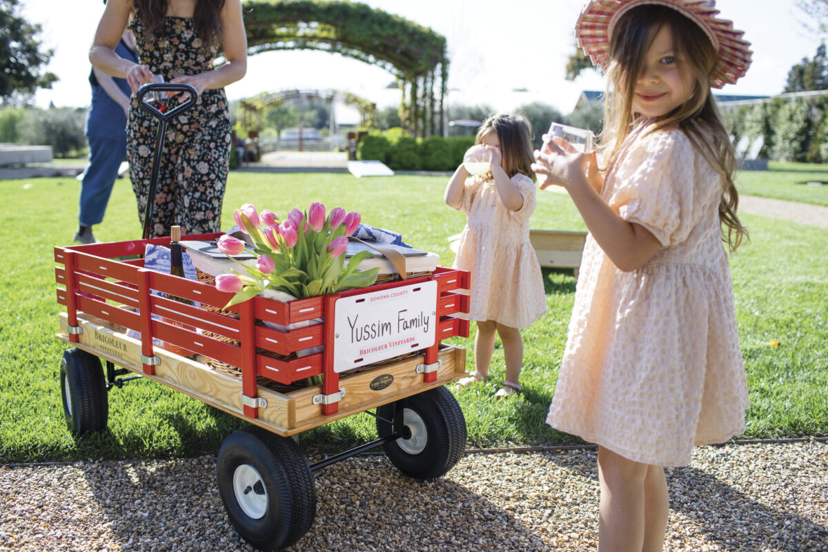 2 girls in dresses with cups of water near a red wagon with tulips in it