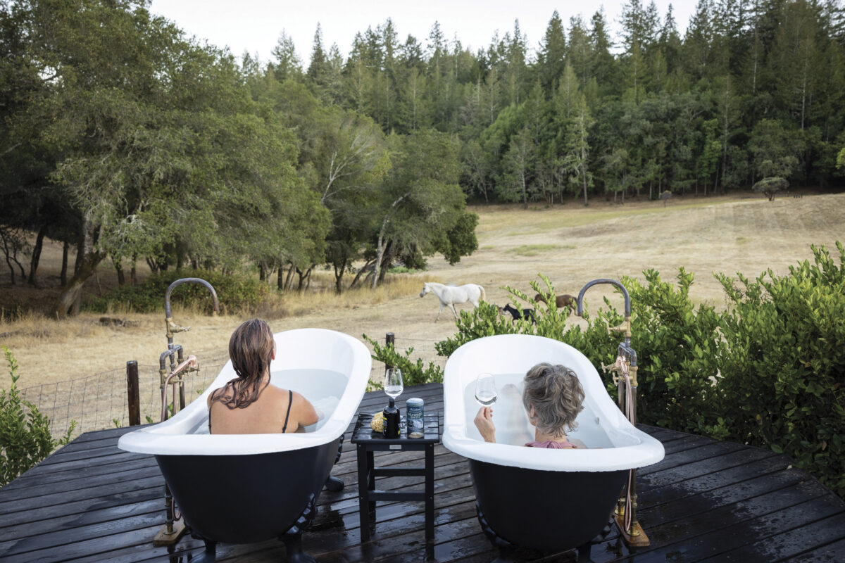 2 women in separate outdoor bathrubs looking out over grazing horses in pasture