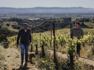 two men and a dog walking in Overshine Vineyards