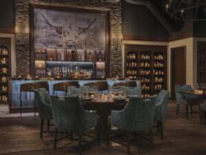 dimly lit, interior image of Ember Steakhouse with wood floors, blue chairs, bar, wine bottles and candlelight