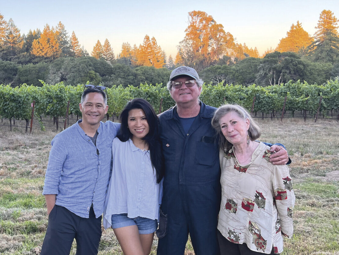 4 adults standing in front of vineyard, smiling with arms around each other