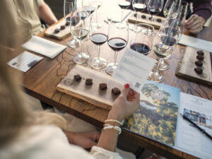 woman holding a tasting menu seated at table with glasses of red wine and wood plate of food