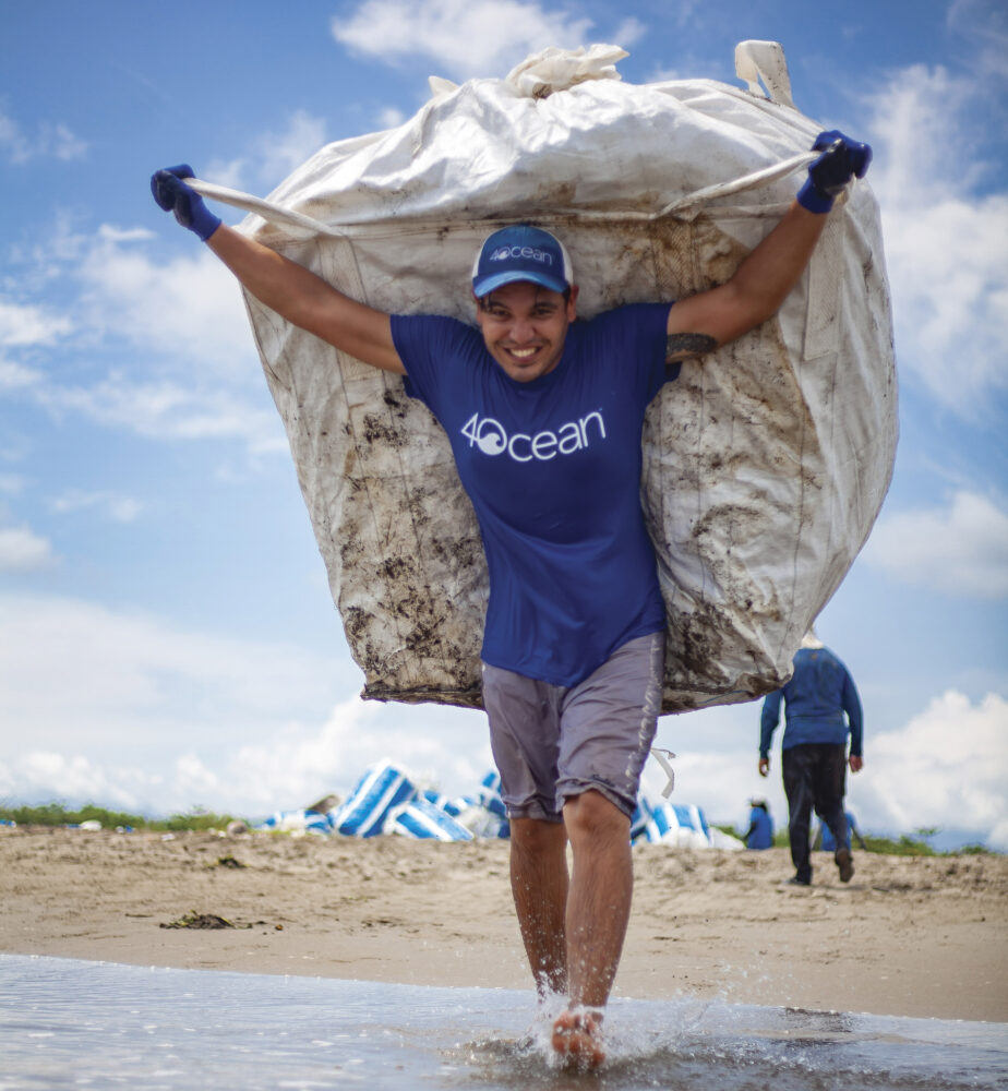 man wearing blue 4Ocean shirt and hat carrying white tarp with gloves walking into ocean water