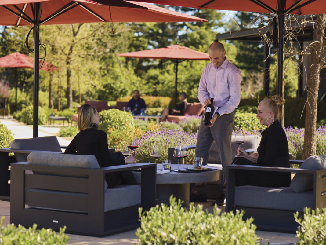 man in dress shirt and pants presenting a bottle of wine to 2 women, seated outside, under red patio umbrellas