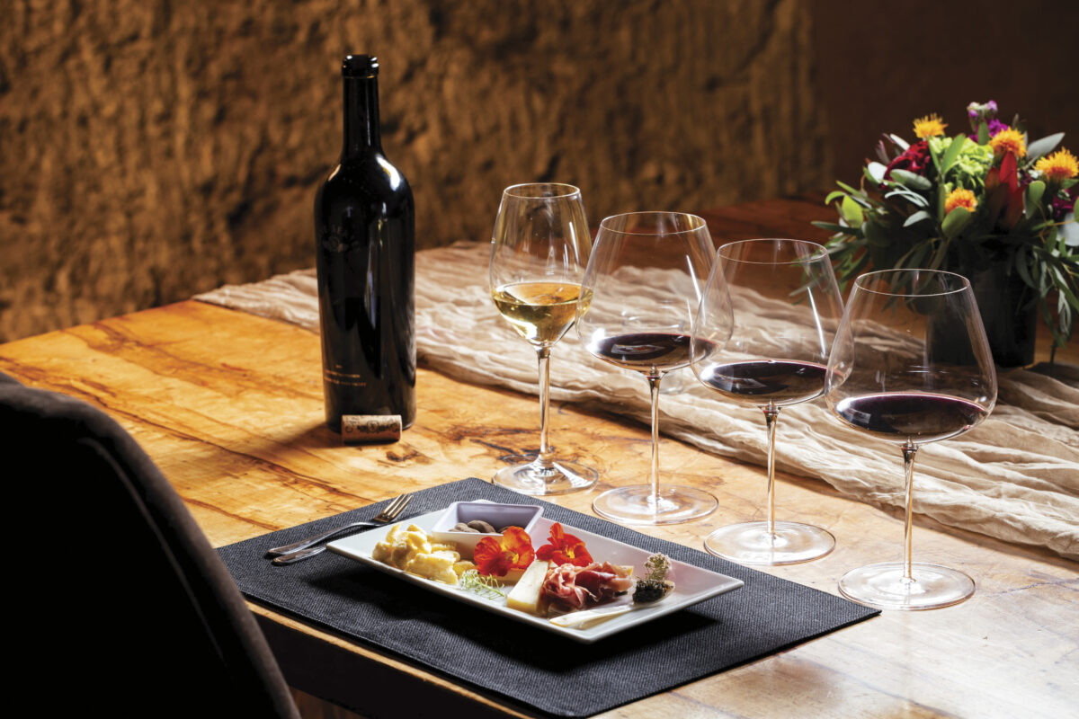 bottle of wine on wood table with 4 glasses set up for wine tasting and plate of food