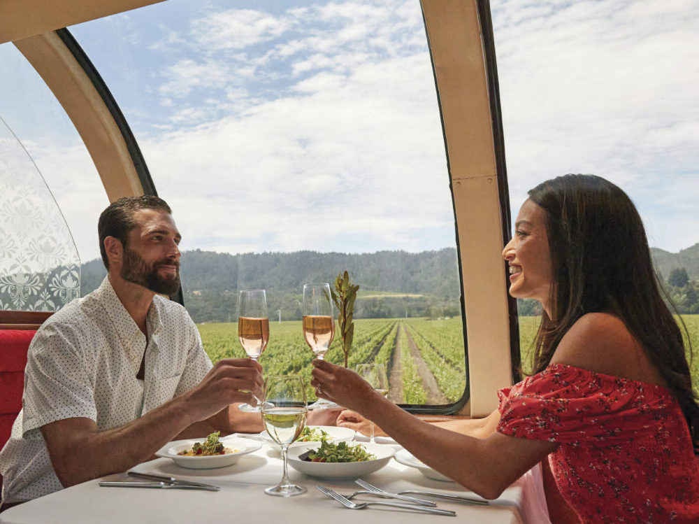 couple tasting champagne flutes, smiling, on train with big window with vineyard and blue sky in the background