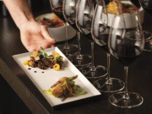 person placing plate of food on wood table in front of glasses of red wine for tasting