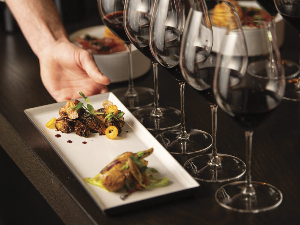 person placing plate of food on wood table in front of glasses of red wine for tasting