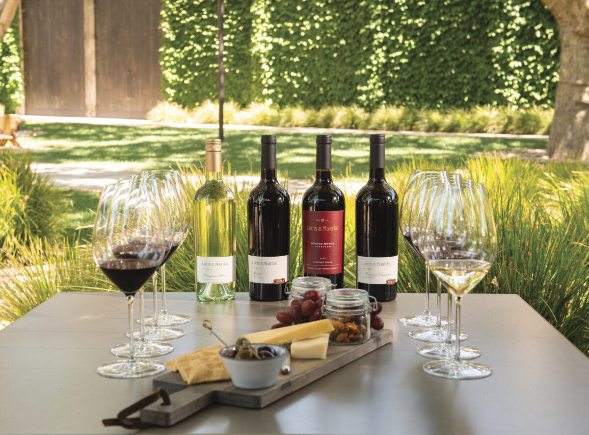 4 bottles of wine, 2 sets of wine glasses set up for tastings with board of cheese and bread set at outdoor table