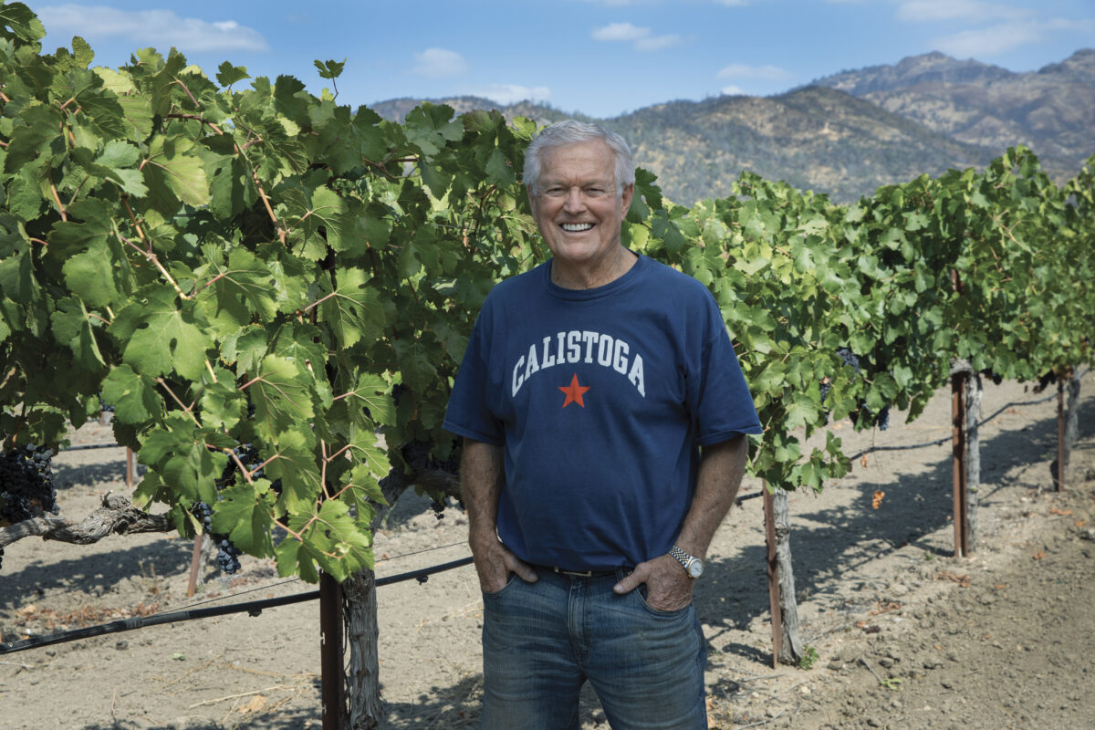 Vermeil proprietor, Dick Vermeil in front of vines wearing a blue tshirt and jeans, smiling