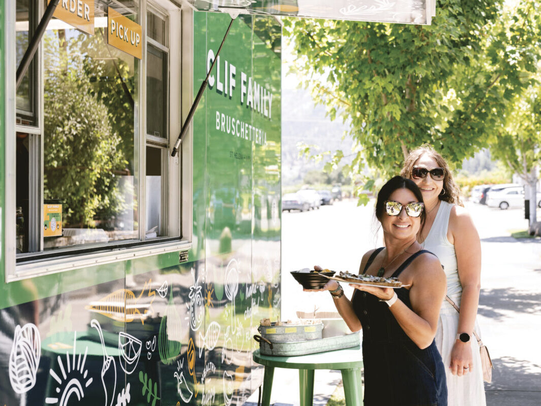 2 women in dresses and sunglasses holding trays of food, smiling, in front of green Clif Family food truck for the article Culinary Crossroads