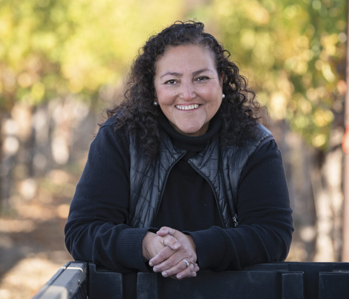 Elizabeth Vianna of Chimney Rock Winery in Napa California wearing black, leaning on bins with vineyard behind her, smiling.