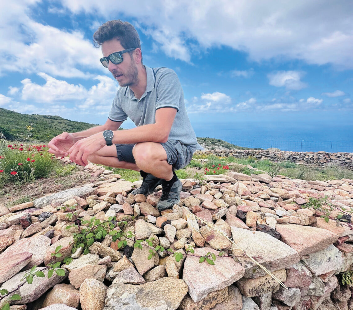 Cosimo Casini wearing shorts and a gray shirt and sunglasses in the Scoglio Nero Vineyard kneeling on rocks with blue sky for the article Heroic Vineyards