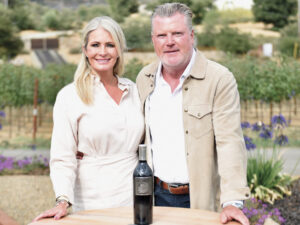 Two adults, Jessica and Chris Zazo, stand in front of vineyard at table with bottle of wine wearing neutral clothing, smiling. For the article Hailstone Vineyards