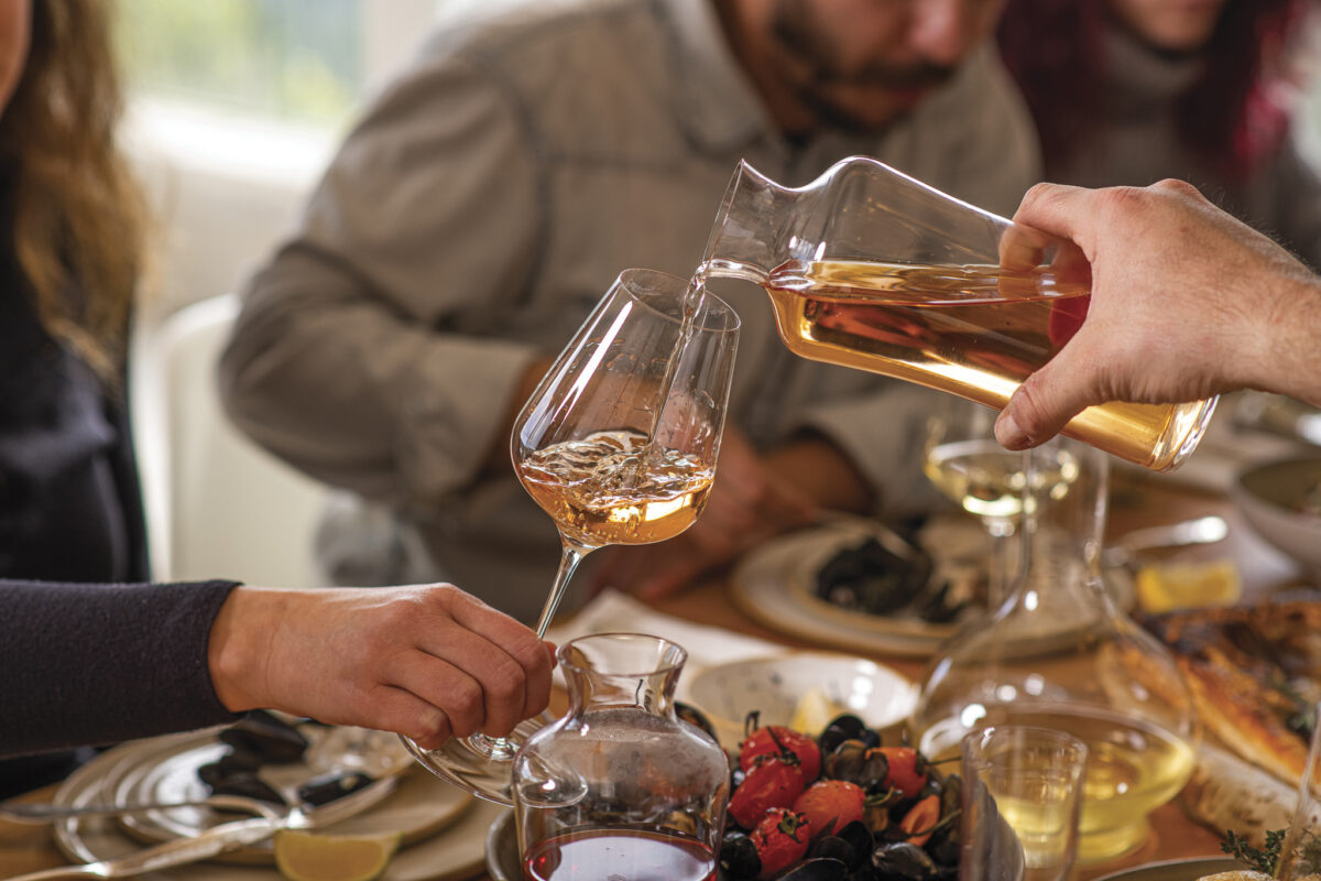 person pouring wine into a glass with others seated at table with wine and food