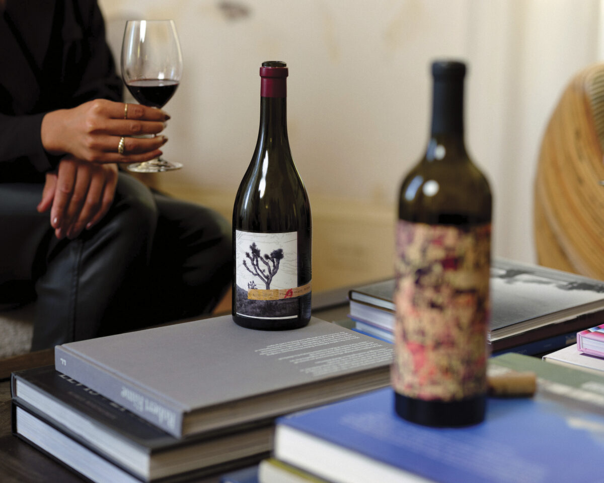 person holding glass of red wine in front of table with stacked books with bottles of wine For the article: Cult Winemaker Dave Phinney