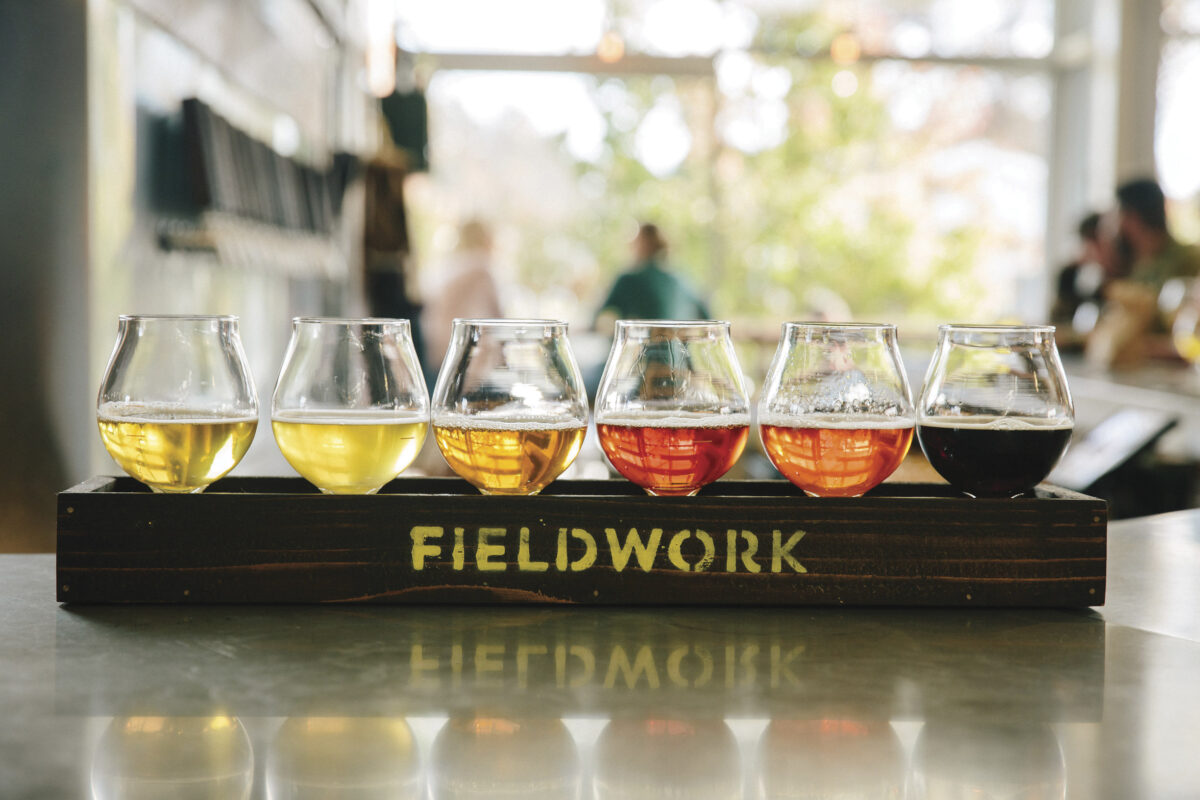 flight of beers is short glasses on wooden tray with company name "Fieldwork" on the side for the article Amuse Bouche