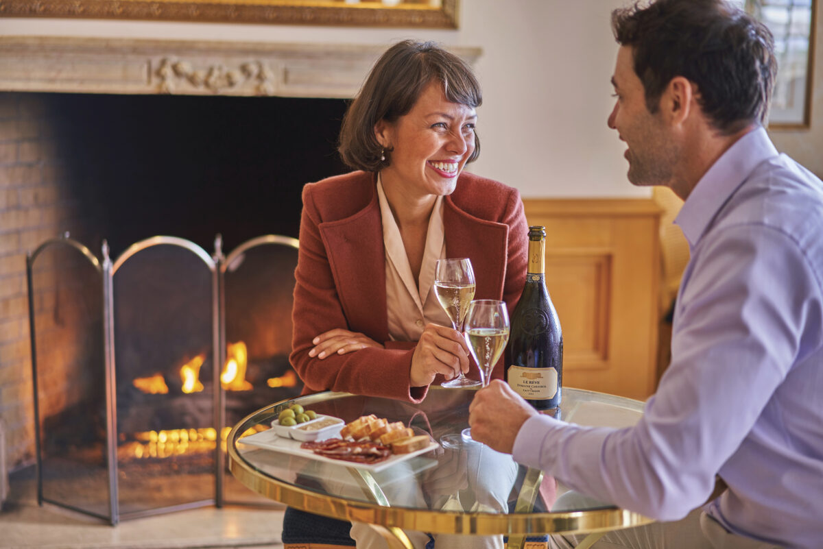 two adults sit at a table with a bottle of bubble and plate of food, indoors near a fireplace holding champagne flutes, smiling for the article Taking Off The Chill 