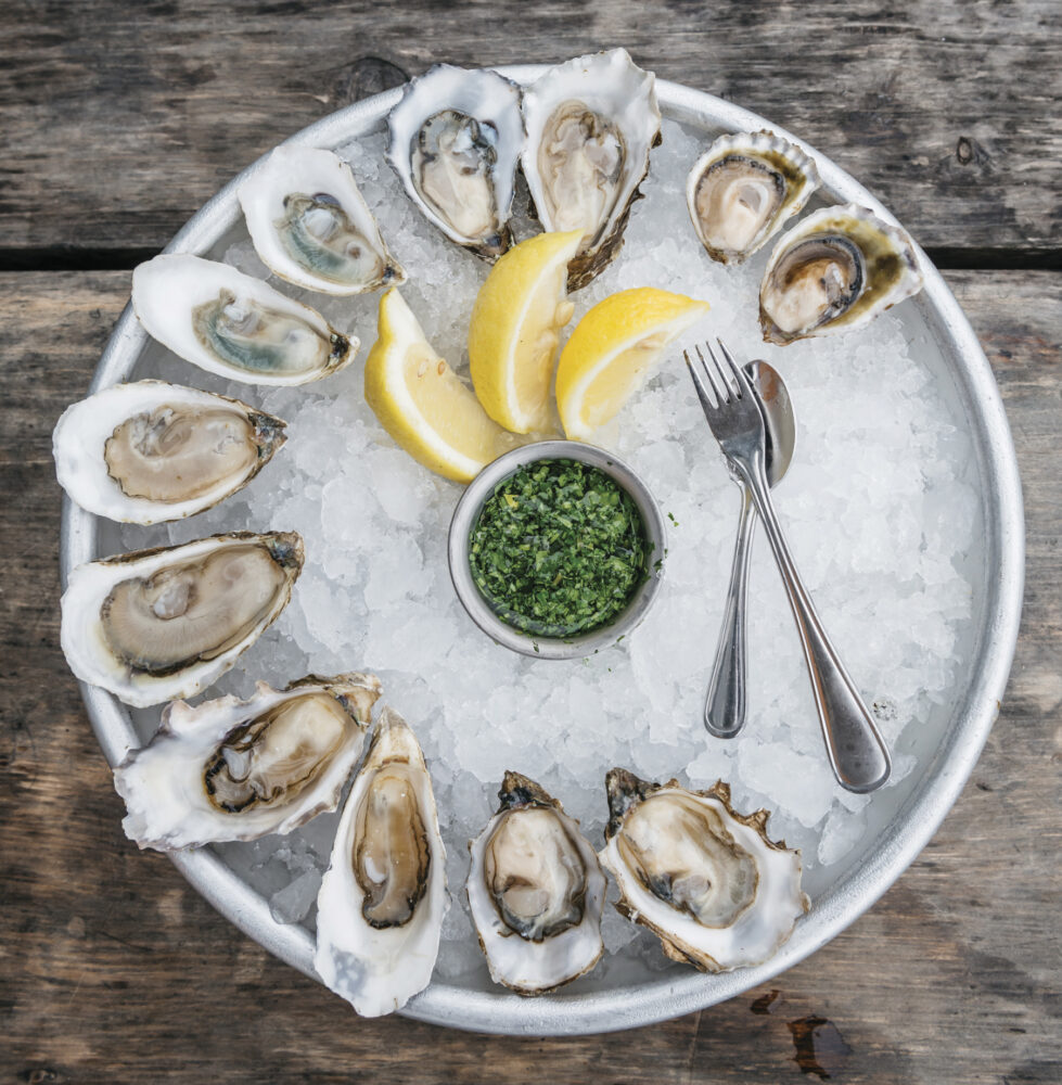 round dish of oysters on ice with slices of lemon, green and utensils on wood table for the article Amuse Bouche