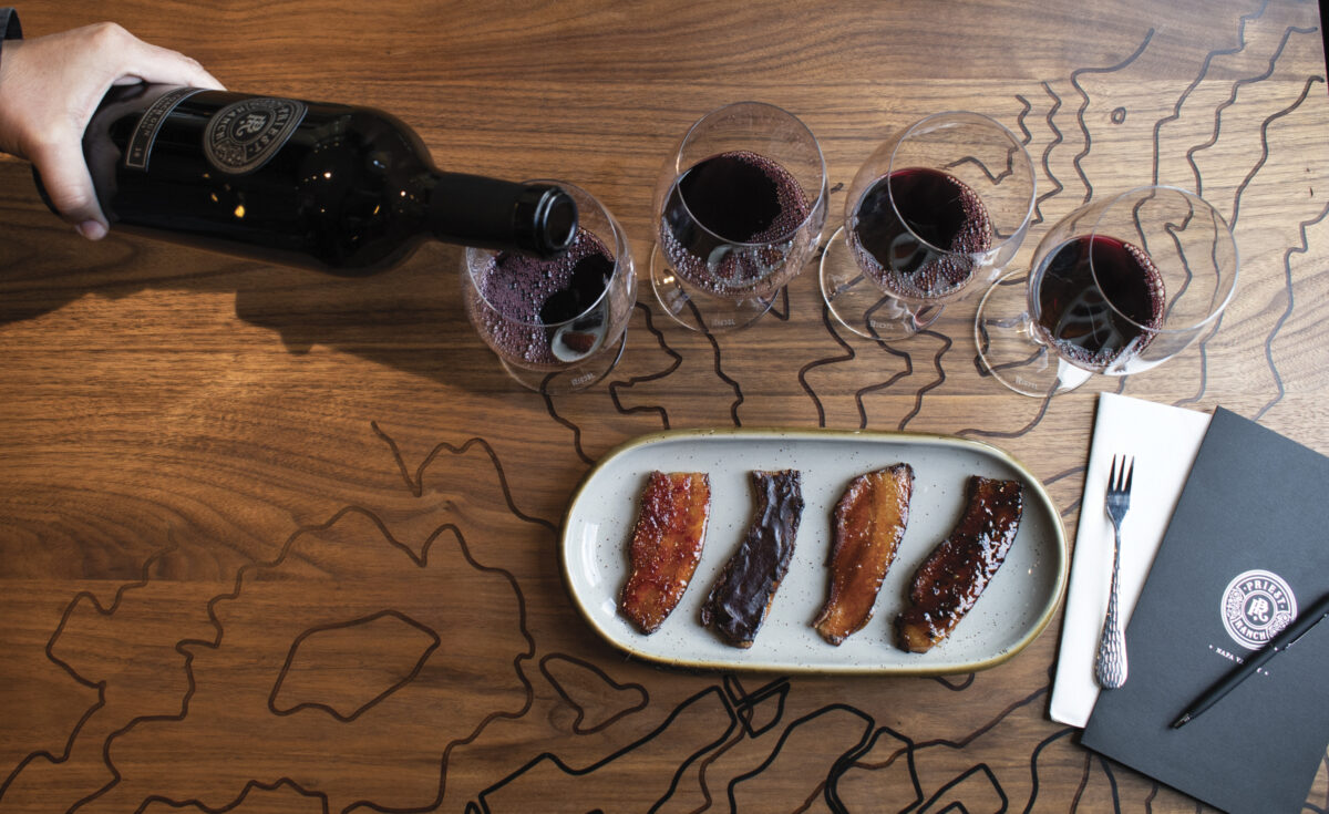 someone pouring red wine into wine glasses on wood table with plate of 4 slices of bacon for tasting for the article "Amuse Bouche"