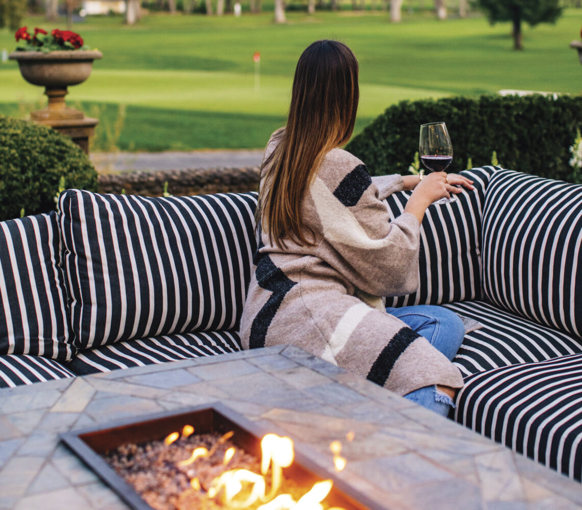 woman seated at outdoor fireplace with glass of red wine facing towards the golf course behind her for the article Taking Off The Chill 