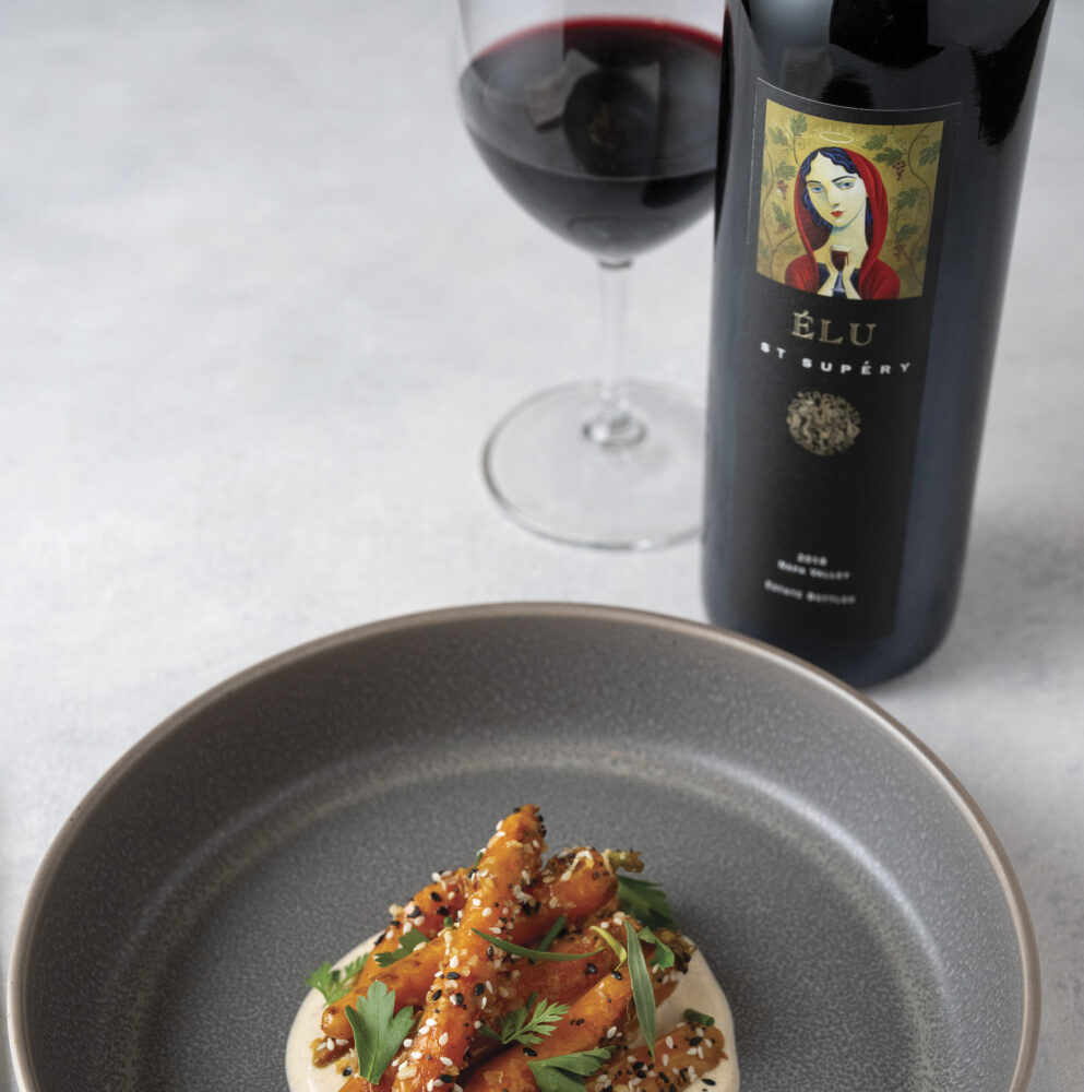 bottle of st. supery wine with red wine in glass with plate of food on gray plate. For the article St Supery Estate Vineyards & Winery