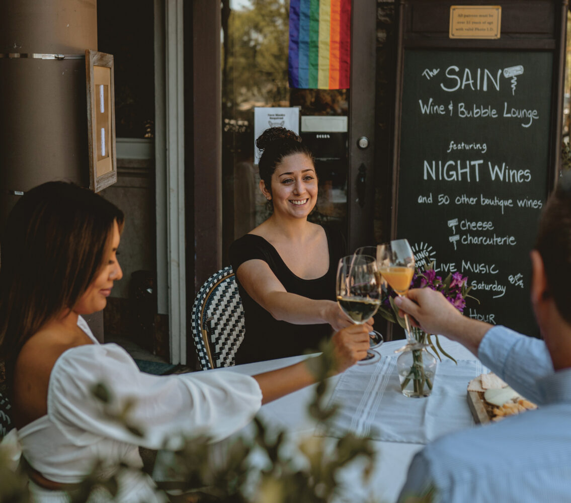 2 women and 1 man seated at outdoor table with white tablecloth, smiling and toasting glassed with rainbow flag hanging above for the article Late Night Napa