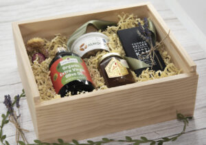 wooden box with a variety of jams and jarred food items on white washed table for article Holiday Gift Guide