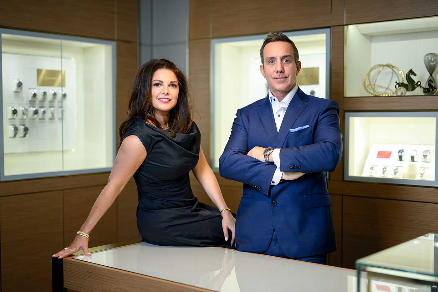 Creations Fine Jewelers owners Melissa (leaning on counter, wearing navy dress, smiling) and Brian Doughty (standing with arm crossed, wearing navy suit) in their newly remodeled store in Napa Valley