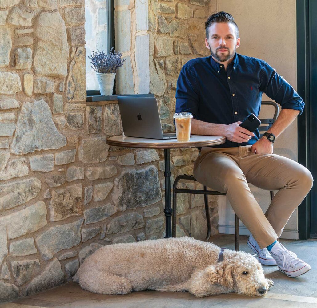 Andrew Allison seated at table with laptop and coffee with dog at feet near stone wall