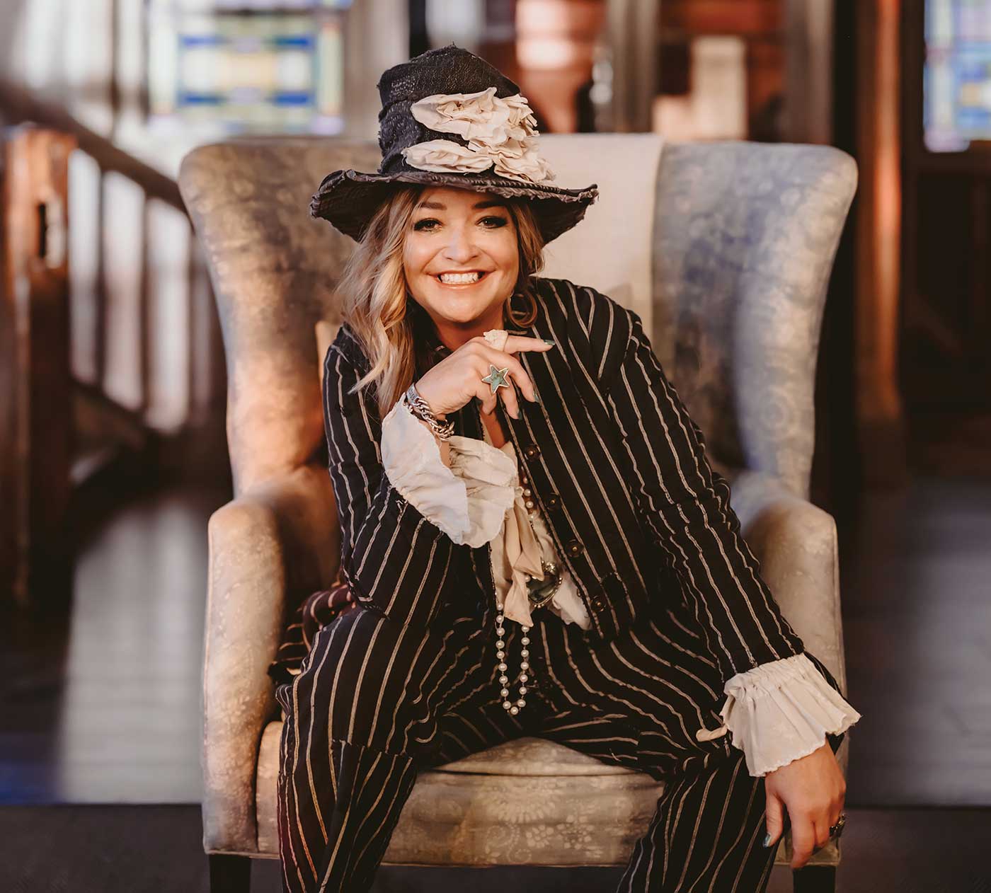 Angela Duerr wearing striped suite with ruffle top and hat, seated in winged chair, smiling for Most Intriguing