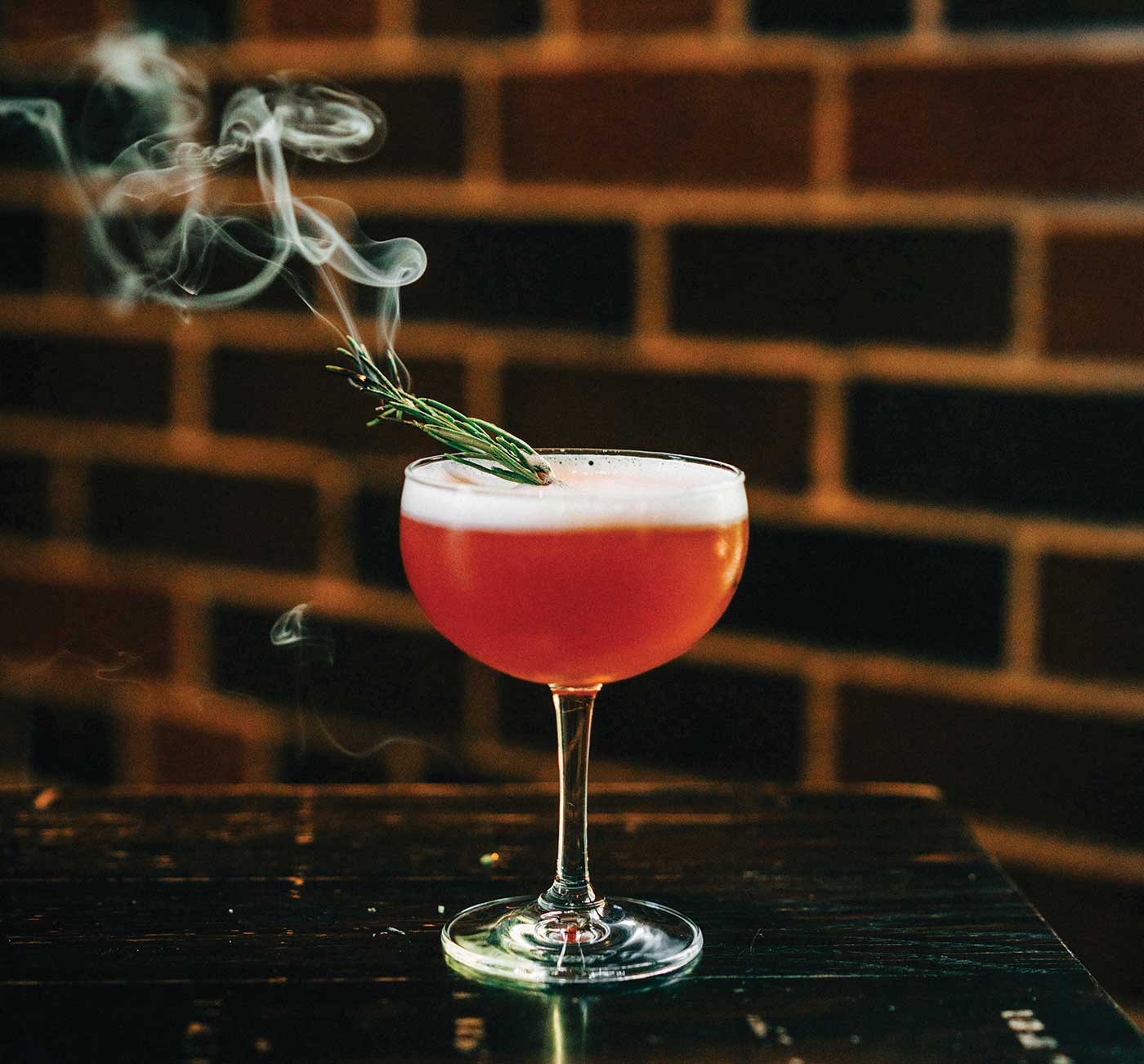 red cocktail with smoke comoing off greenery in cocktail glass on wood table in front of brick wall - Charter Oak's Crimson Bloom cocktail for seasonal sipping