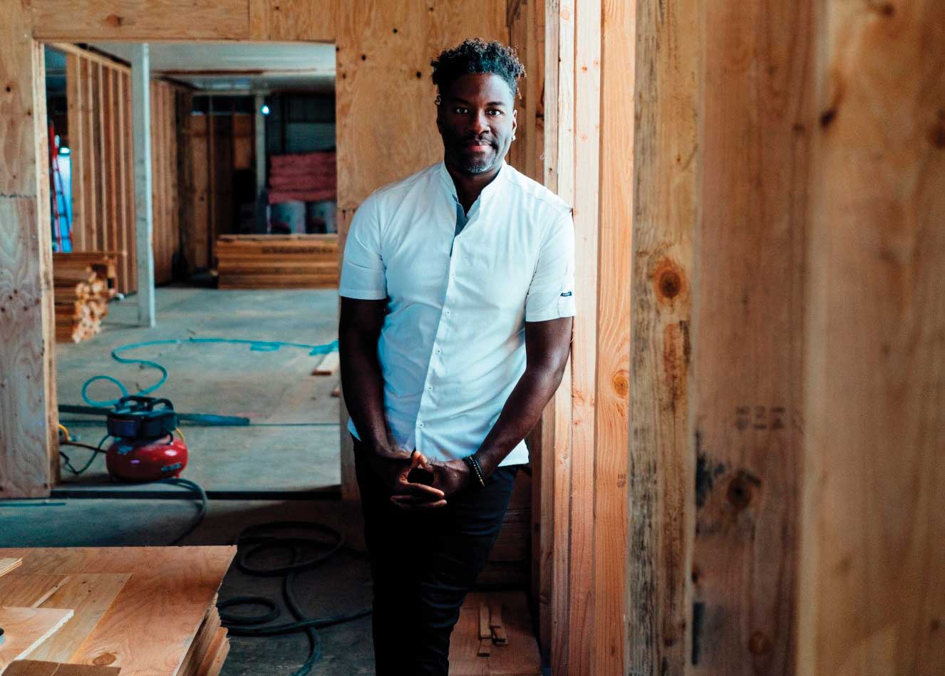 Darryl Bell leaning on wooden stud wall in white shirt and black jeans in rom that's under construction 