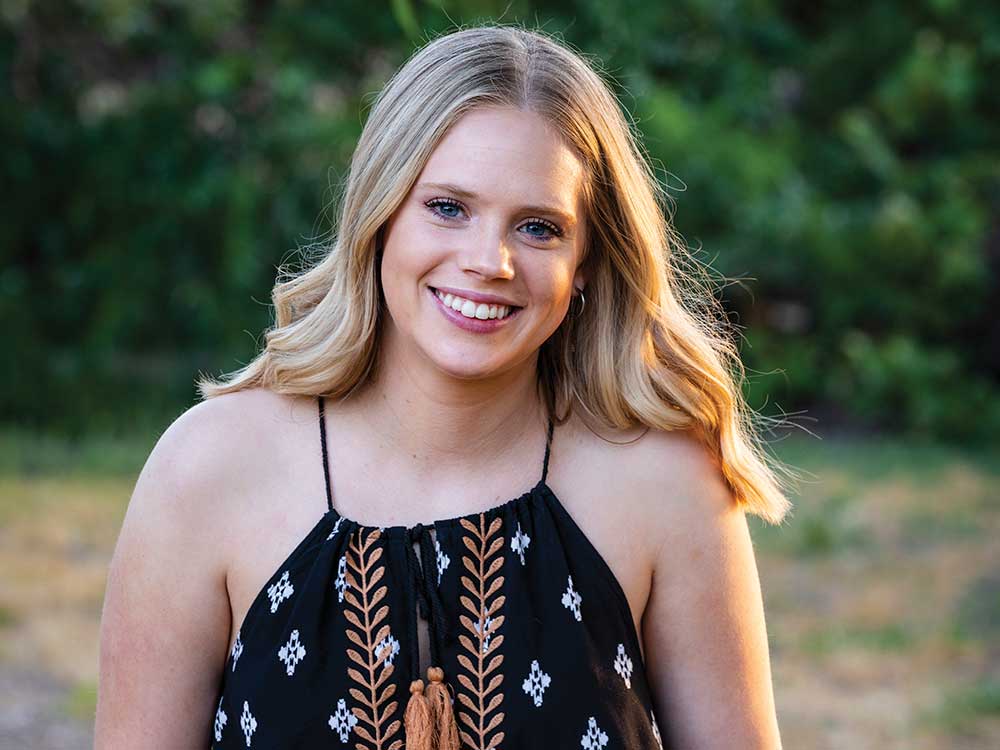 Elizabeth Shaw, abstract artist, posing in black top, smiling with greenery behind her