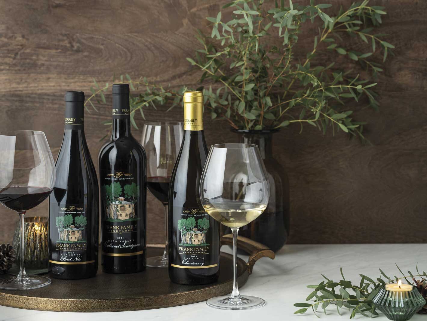 Assortment of Frank Family Vineyards wine bottles and glasses of red and white wine on white table with wooden tray and greenery with wood background for Solar sustainability