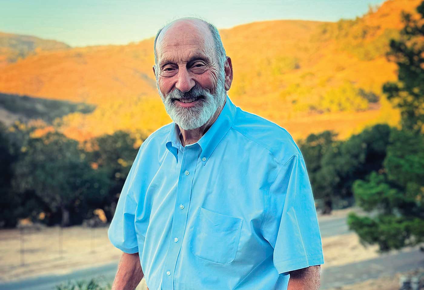 John Tuteur standing outside with mountain and trees behind him, smiling wearing light blue shirt