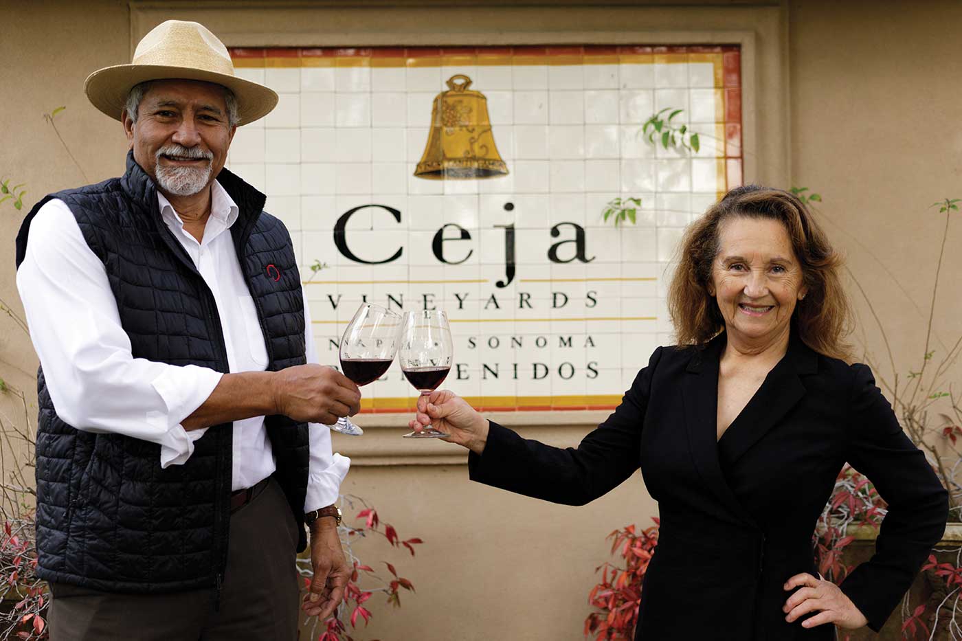 Pedro and Amelia Ceja toasting red wine glasses, smiling in front of the Ceja vineyards sign 