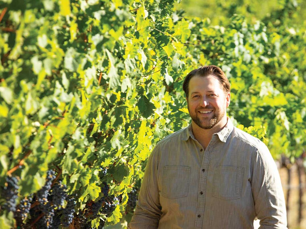 Chris Kenefick standing in front of vineyard wearing khaki shirt, smiling in Calistoga